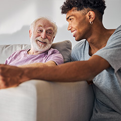 a carer kneeling next to a sofa and speaking to a smiling elderly man