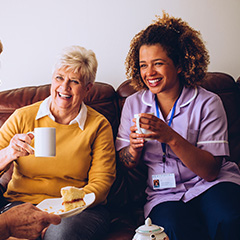 a carer smiling next to an elderly lady while the both share a cup of tea together